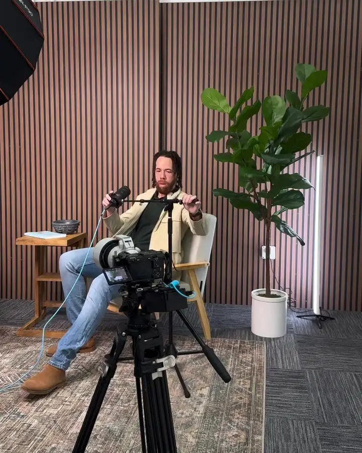 Podcast set with a host in an armchair, a camera on a tripod, a plant, and wood slat walls