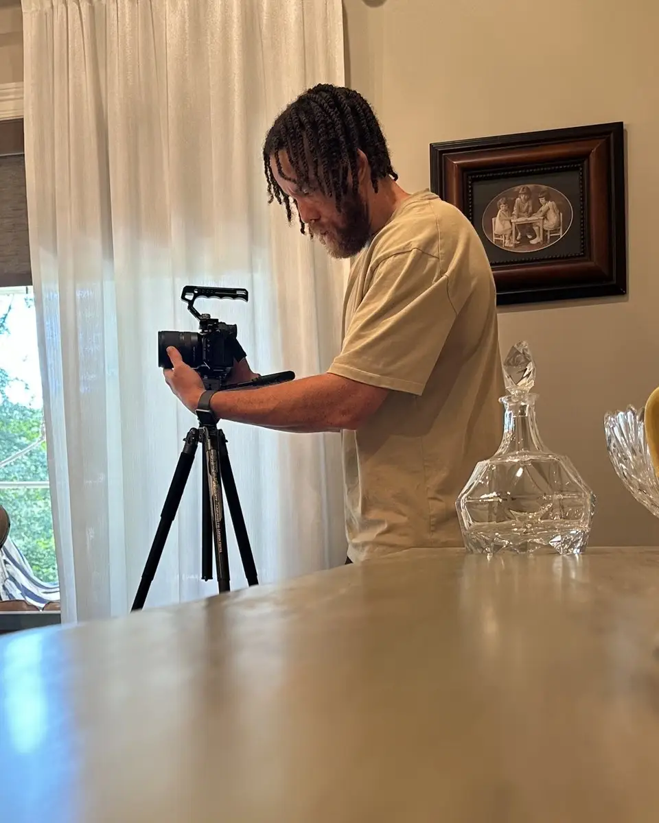 Tyler Thompson filming with a camera on a tripod inside a home, sheer curtains behind him